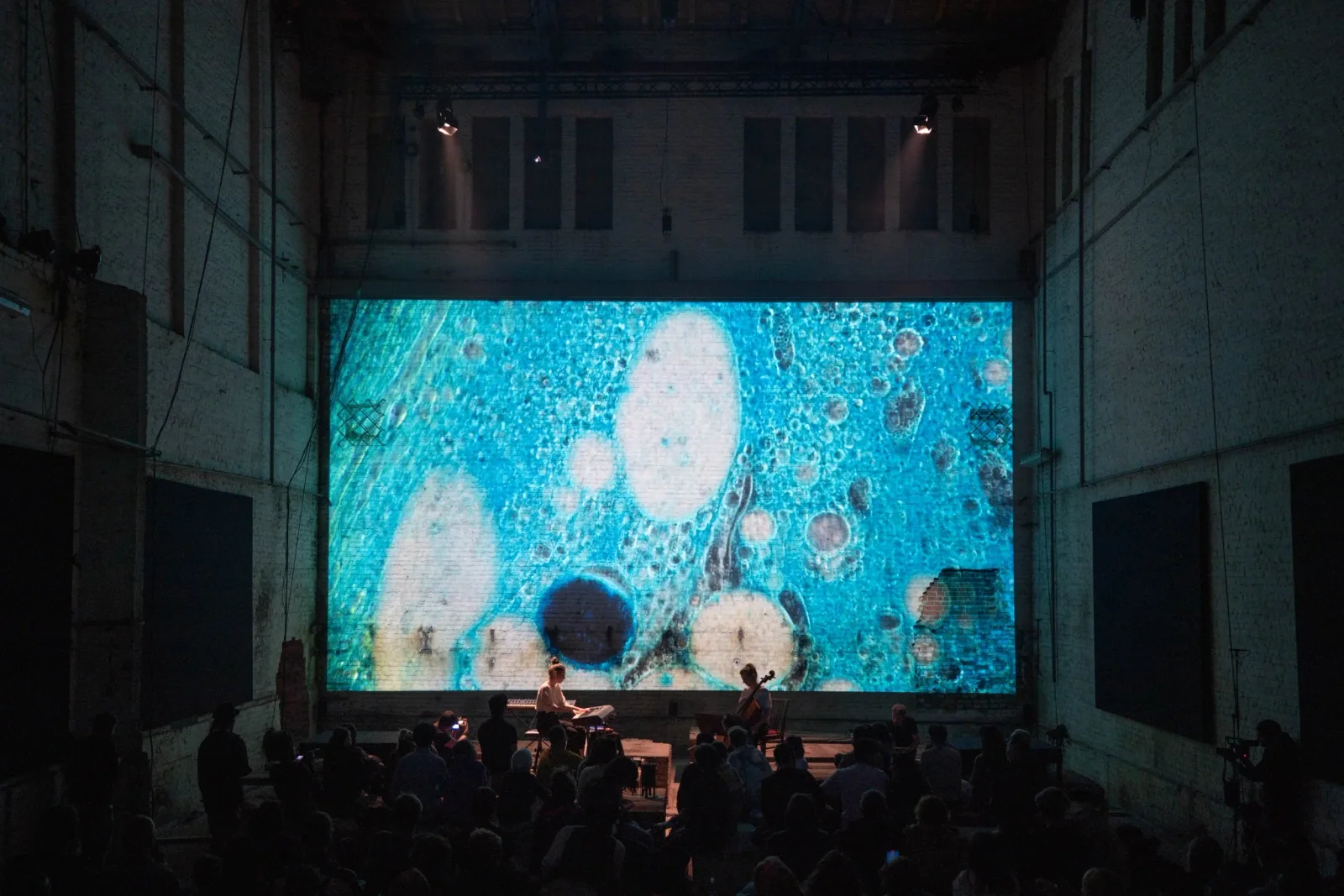 Wide audience view of a pianist and cellist performing beneath large blue microscopic projections in the Monopol hall.