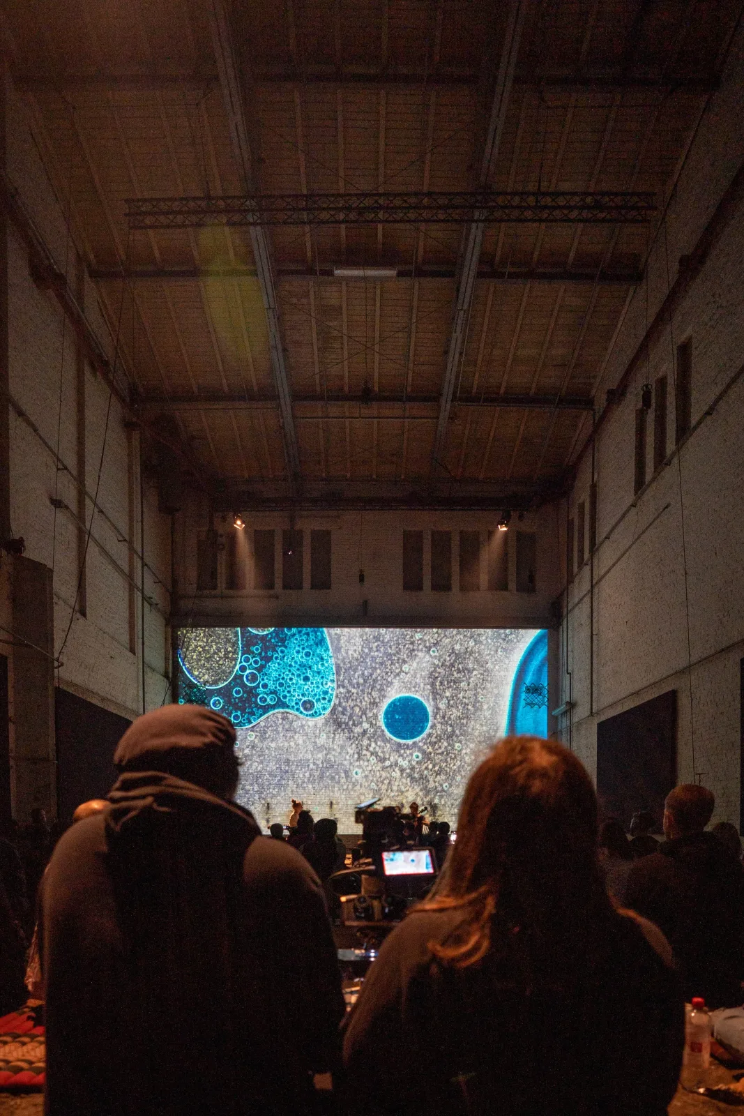 Tall view through the audience toward the stage, production camera and blue microscopic projection inside Monopol.
