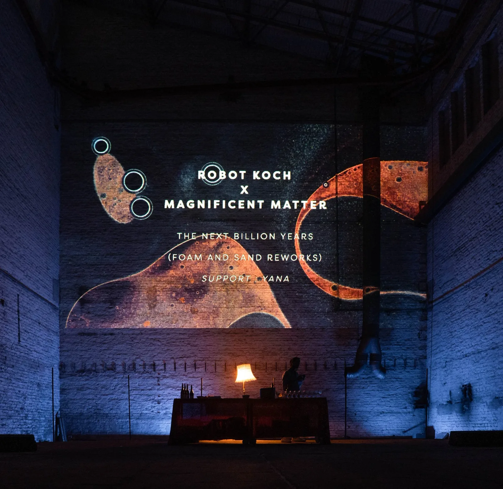 Event title projected in white over orange microscopic shapes on the brick wall at Monopol.