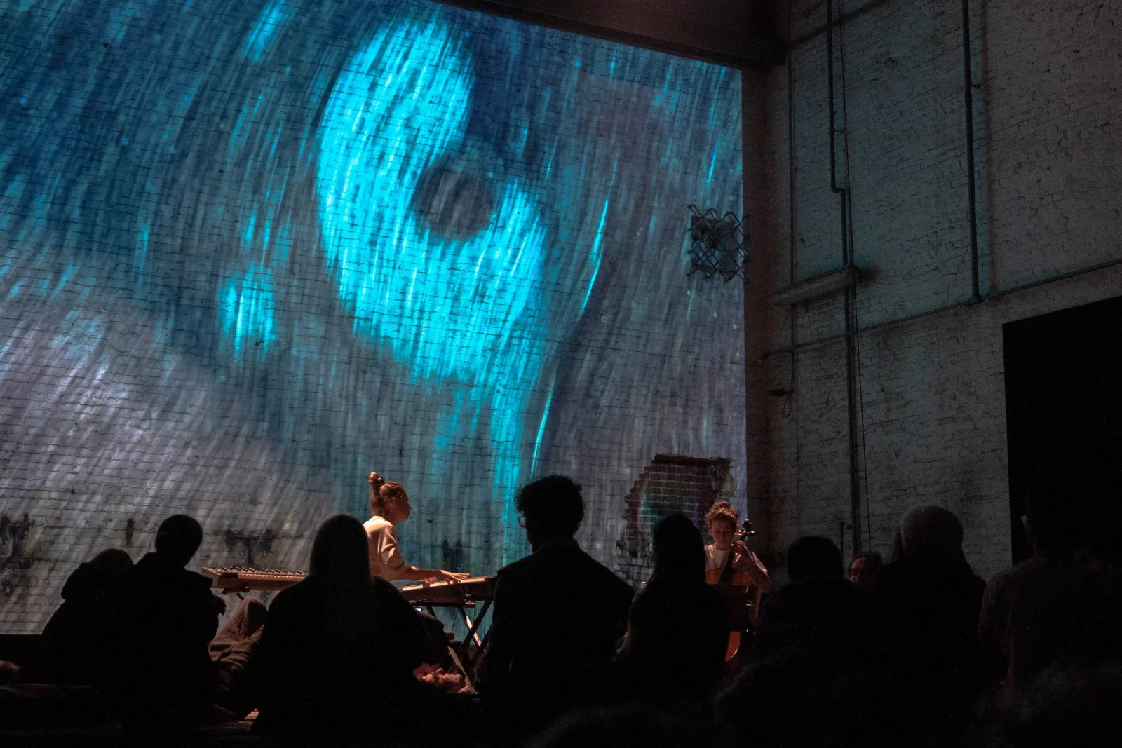YANA and Maria Szagżdowicz perform before an audience while blue abstract imagery spans the brick wall behind them.