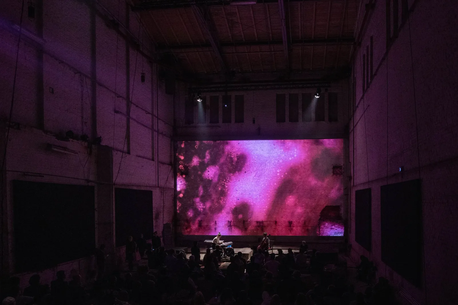 Pianist and cellist on stage beneath a large magenta microscopic projection, viewed across the audience.